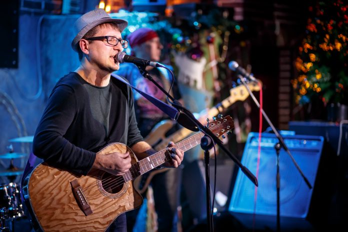 Musician wearing a hat and glasses, playing an acoustic guitar and singing into a microphone during a live performance, with colourful stage lights in the background