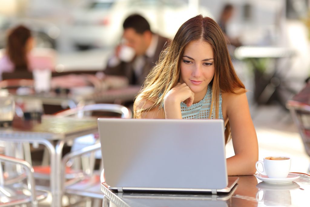 Young woman working on a laptop at an outdoor café, representing the lifestyle of a freelance coder