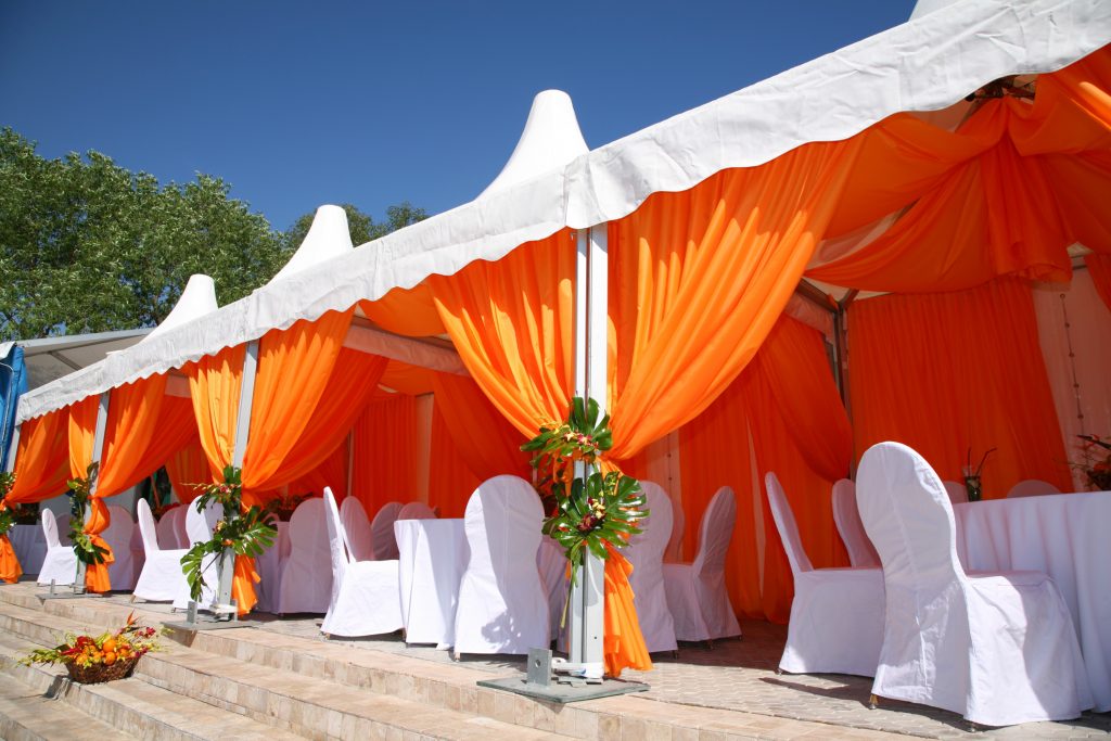 Beautifully decorated outdoor event tent with vibrant orange drapes and white-covered chairs, exemplifying the creativity of an event planner