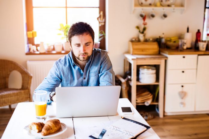 Man working remotely from home on a laptop with a cup of orange juice and a croissant on the table