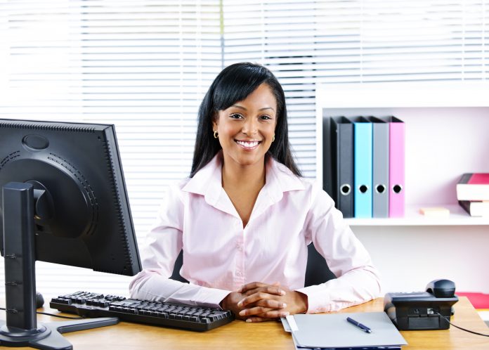 Professional woman in a light pink blouse smiling at her desk in a bright, modern office with a computer monitor and organized shelves in the background