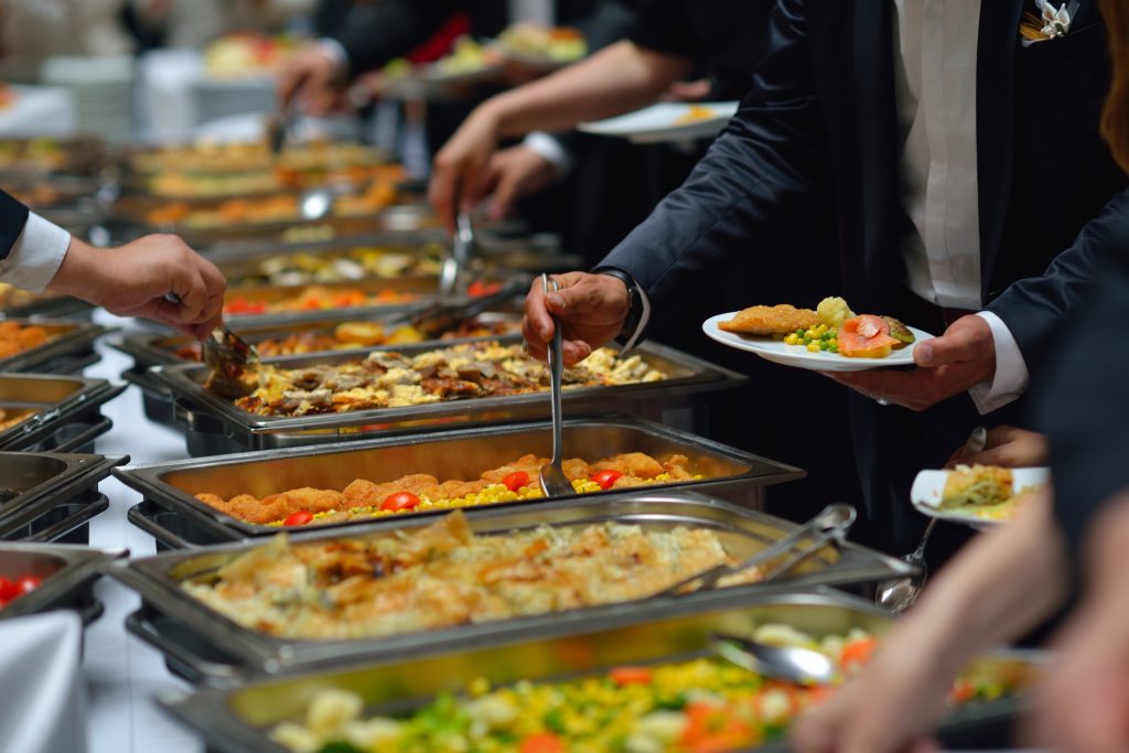 People serving themselves food from a buffet table filled with various dishes in stainless steel trays at a catering event