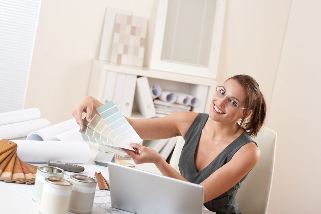 Interior designer holding and reviewing colour swatches at her desk, surrounded by design materials such as paint cans, wood samples, and architectural drawings