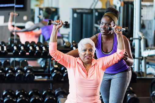 Personal trainer assisting an older woman with a shoulder press exercise in a gym, emphasising personalised fitness support